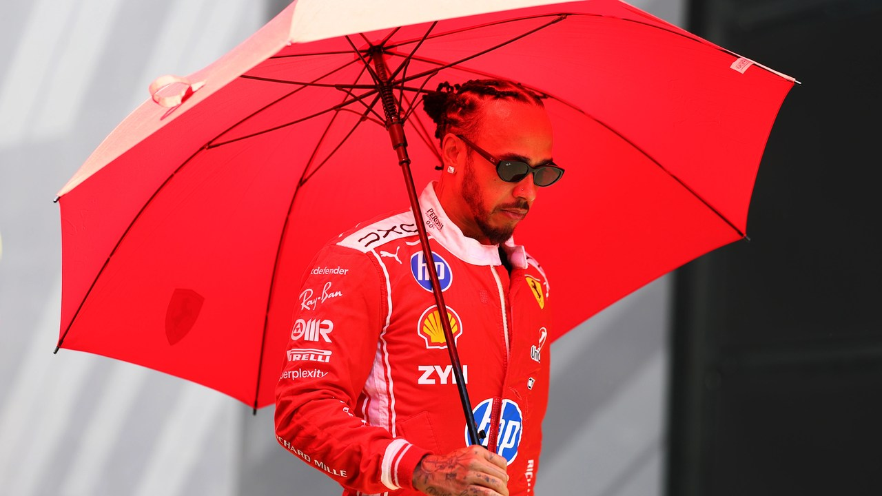 Lewis Hamilton, Ferrari takım kıyafetleriyle kırmızı bir şemsiye altında yürüyor.